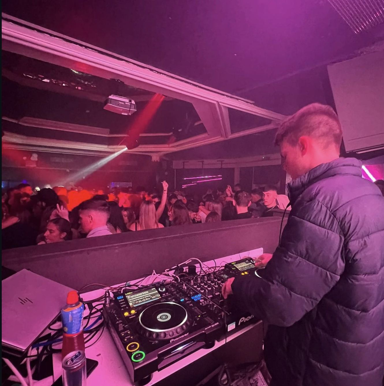 George in the DJ booth playing to a packed club dancefloor under pink and red lights