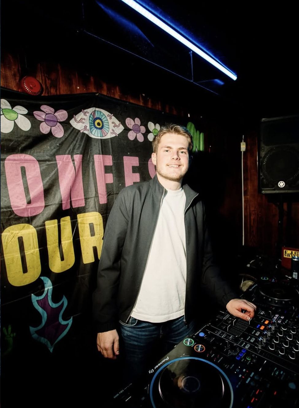 George smiling behind the decks, with a colourful banner behind him